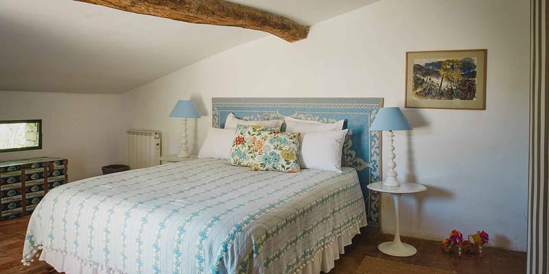 An interior shot of a guest bedroom at what appears to be a French country property, likely used as accommodation for a wedding event. The room features a double bed with a blue-and-white tiled or embroidered headboard, dressed in a white and light blue floral quilted bedspread with pom-pom trim and multiple white pillows, including a multicolored floral accent cushion in blue, yellow, green, and red. Two matching white candlestick-style lamps with pale blue shades sit on either side of the bed on white side tables. The room has a vaulted ceiling with an exposed rustic wooden beam, terracotta tile flooring, and white walls with a framed artwork print above the right nightstand. A vintage trunk is visible in the background left corner. The overall styling is rustic-provincial with blue and white tones. Wide interior shot. Potential venue feature image.