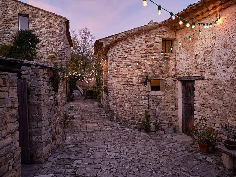 Rustic stone alleyway at dusk with globe string lights strung between buildings and flagstone pathway