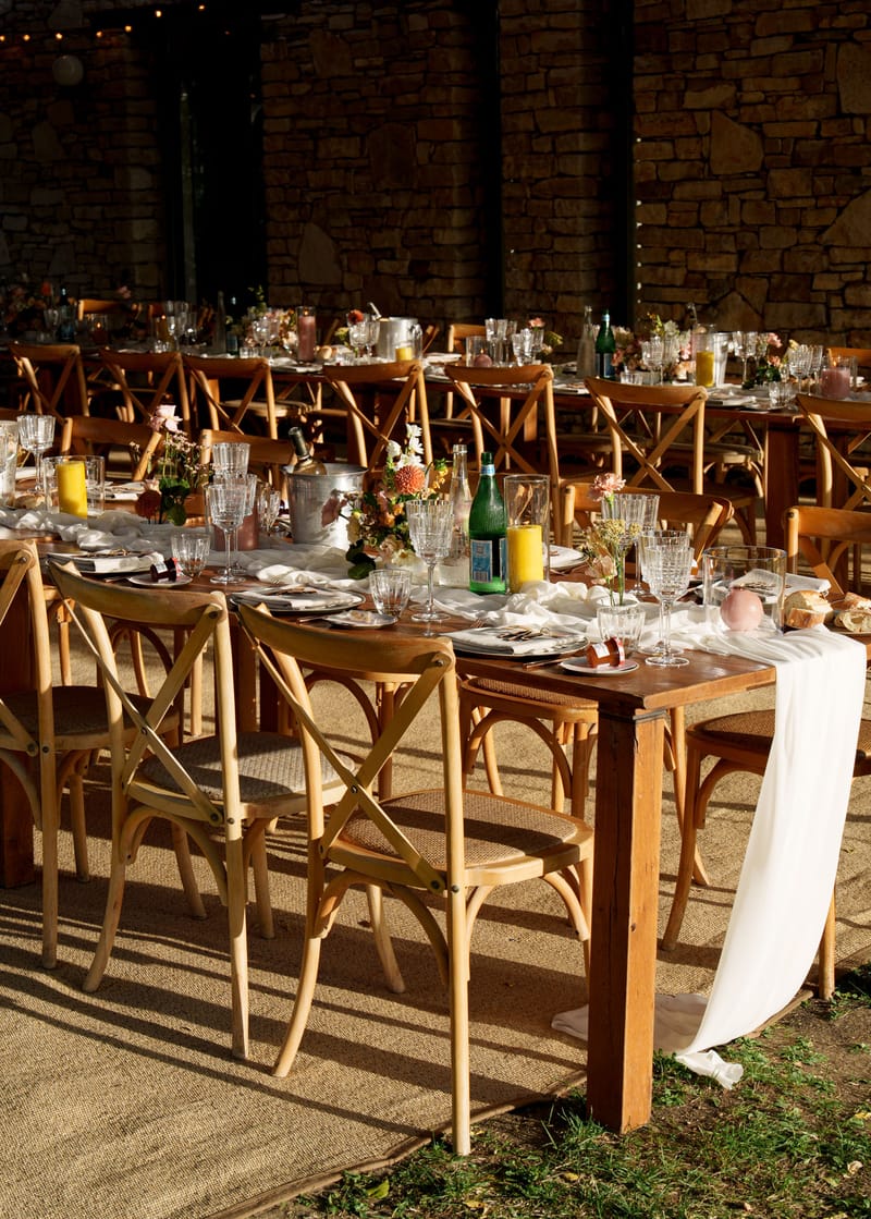 A wedding reception tablescape set under a covered outdoor space with exposed stone walls and string lights overhead. Multiple long wooden farm tables are arranged with natural wood cross-back chairs and laid with white linen runners draping over the edges. Each place setting includes charger plates, folded white napkins, and cut-crystal glassware. Centerpieces feature low arrangements of coral and orange dahlias, pink blooms, and small bud vases with wildflowers, alongside yellow pillar candles, wine bottles in silver ice buckets, and green glass water bottles. The overall decor palette combines warm terracotta, amber, and dusty rose tones with natural wood and white linen, creating a rustic yet refined aesthetic. Wide shot capturing the full table layout from a slightly elevated angle.