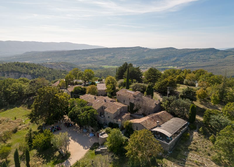 An aerial drone shot captures a large Provençal stone farmhouse complex with terracotta-tiled roofs set among dense trees, with a wide valley and mountain range visible in the background. The property comprises several interconnected stone buildings arranged around a gravel courtyard, with a covered barn structure visible to the right and a swimming pool visible further back on the grounds. A group of approximately 30–40 wedding guests can be seen gathered in the courtyard to the left of the main building, suggesting a ceremony or cocktail hour in progress outdoors. The wide establishing shot showcases the full scale of the venue, its rural Provençal architecture, and its elevated hillside position. Potential venue feature image.