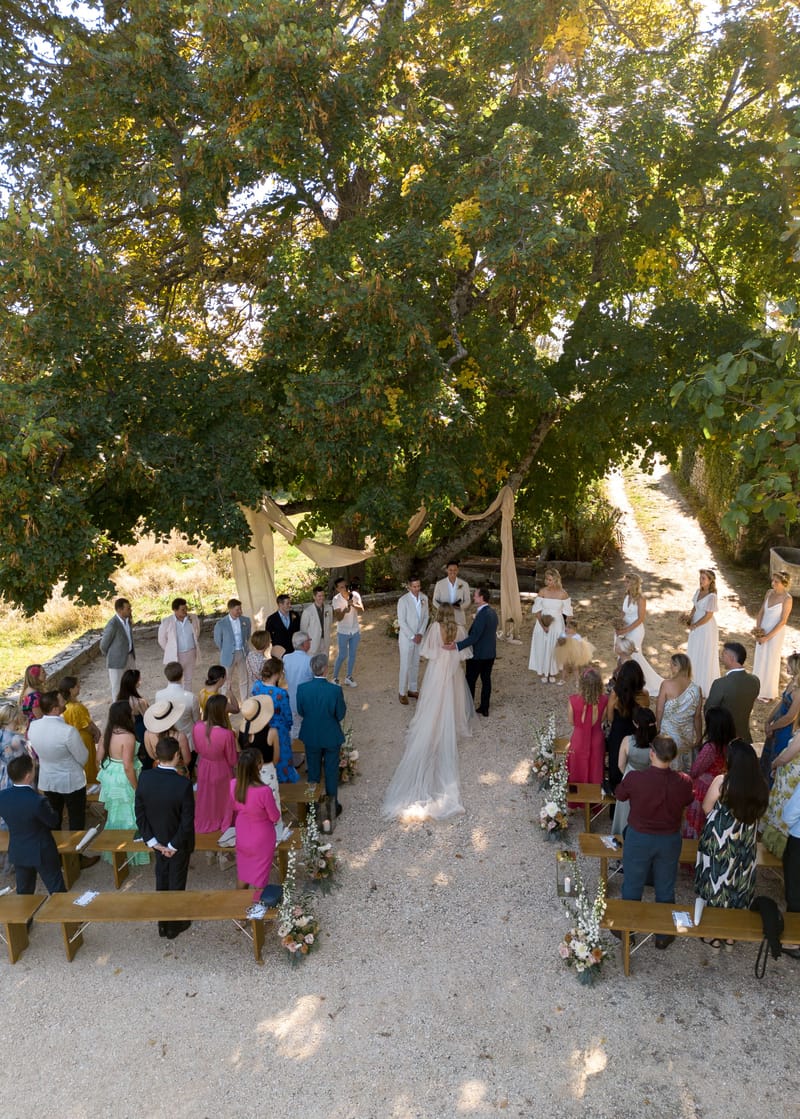 An outdoor wedding ceremony photographed from an elevated angle, showing the couple standing at the altar beneath large mature trees on a gravel surface. The bride wears a blush-pink gown with a long veil, and the groom is dressed in a navy suit; they face an officiant who is reading from a book. The altar area is framed by draped natural linen fabric tied loosely between tree branches, with arrangements of white delphiniums, blush roses, and foliage placed along the aisle on wooden bench ends. Approximately 40–50 guests are seated and standing on wooden benches arranged in two sections, dressed in a mix of colorful outfits including fuchsia, cobalt blue, mustard, and mint. The bridal party on the right side of the altar wears white dresses, while the groomsmen on the left wear a mix of light linen and grey suits. The overall styling is relaxed boho with natural, earthy tones in the decor.
