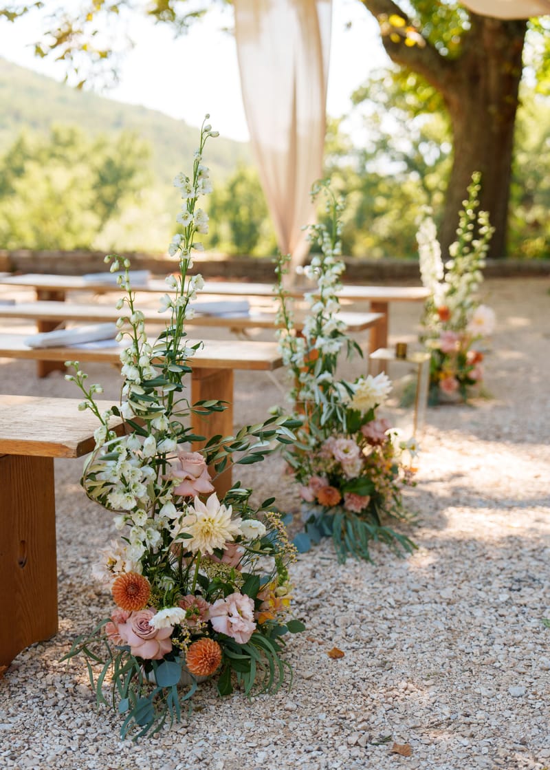 Ceremony aisle floral arrangements of cream dahlias, dusty rose roses, and orange blooms beside wooden benches