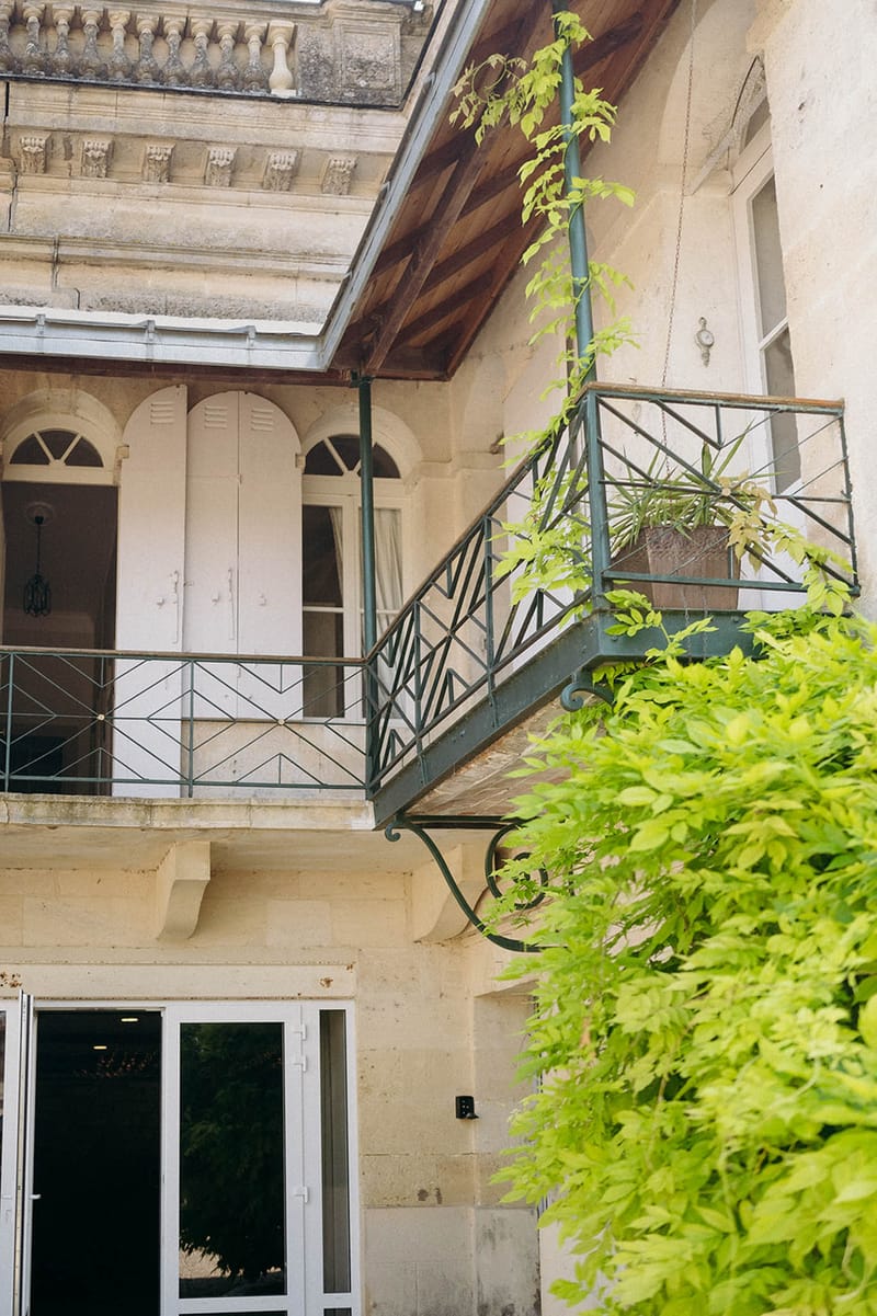 An exterior architectural shot of a French manor or château-style building, showing the facade of an interior courtyard. The cream-colored limestone building features two levels of dark green wrought-iron balconies with geometric chevron-patterned railings, arched windows with white shutters, decorative carved cornices, and exposed wooden beam roofing visible at the top. A climbing vine grows up one of the balcony supports, and a potted plant sits on the upper right balcony. Ground-floor double glass doors are visible at the bottom left. No people are present in the image. Wide shot, portrait orientation. Potential venue feature image.