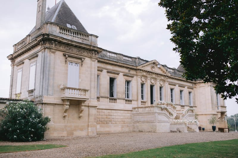 A wide exterior shot of a French château with classic 18th-century architecture, featuring cream limestone façades, ornate carved stonework, tall shuttered windows, a balustraded terrace, and a pointed slate-roofed tower on the left. The entrance staircase is decorated with arrangements of white flowers — appearing to be white roses and possibly baby's breath — draped along the balustrade and flanked by potted topiaries in dark planters, indicating the venue is dressed for a wedding. No people are visible in the frame. The gravel forecourt and manicured lawn are visible in the foreground. Potential venue feature image.