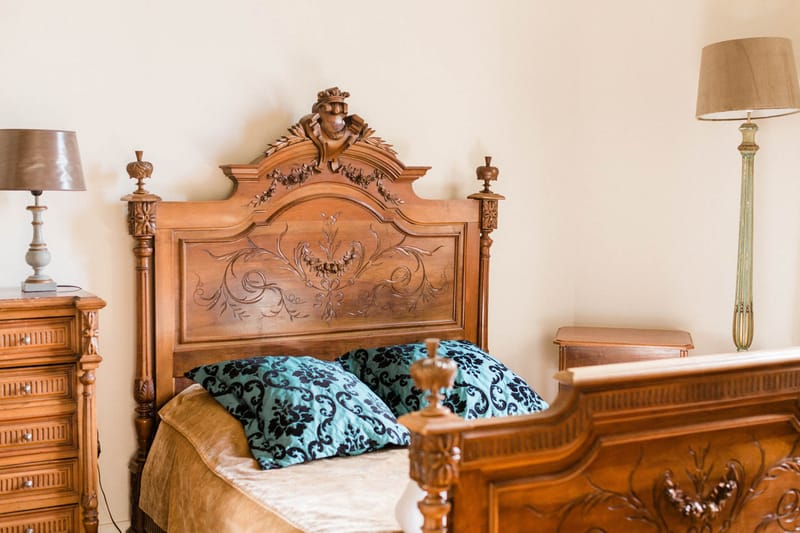 An interior shot of a bedroom, likely within a château or manor house used as a wedding venue. The room features an ornate antique walnut bed frame with deeply carved floral and scrollwork motifs on the headboard, topped with a carved figurehead at the crest. The bed is dressed in a caramel-toned bedspread with two teal velvet cushions printed with a black damask pattern. The matching carved walnut bedside chest and footboard are visible, flanked by two table lamps — one with a bronze-toned shade on the left and a tall floor lamp with a taupe shade on the right. The overall aesthetic is classic French antique, with warm wood tones against cream walls. Wide interior shot. Potential venue feature image.
