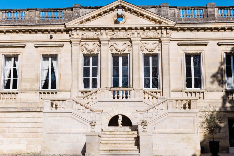 A wide exterior shot of a French classical château façade built in pale limestone. The building features a central pediment with decorative carved stonework including garland and wreath motifs, tall white-framed French windows across two visible floors, and a grand double staircase with balustraded railings leading up to a landing. A small classical stone statue is visible beneath an arched niche at the base of the staircase, and ornamental urns flank the steps. No people are visible in the image. The photograph is taken straight-on in bright daylight with no wedding-specific decor present. Potential venue feature image.