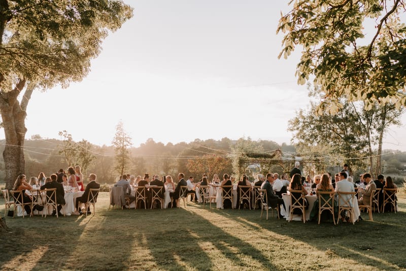 An outdoor wedding reception dinner is underway on a large lawn, with approximately 60–80 guests seated at multiple round tables covered in white linens. The tables are surrounded by natural wood cross-back chairs, consistent with a rustic-country styling theme. String lights are strung overhead between the trees, and a greenery-covered pergola structure is visible in the background center, draped with climbing foliage. The scene is lit by warm late-afternoon golden hour light, casting long shadows across the lawn and bathing the entire setting in an amber glow. Guests are dressed in a mix of formal and semi-formal attire, including dark suits and dresses in black, red, and neutral tones. The wide-angle shot is taken from ground level looking across the lawn toward the open countryside beyond.