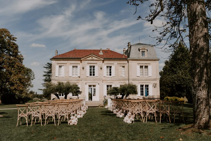 Outdoor ceremony setup with wooden cross-back chairs and blush floral aisle markers facing a limestone chateau