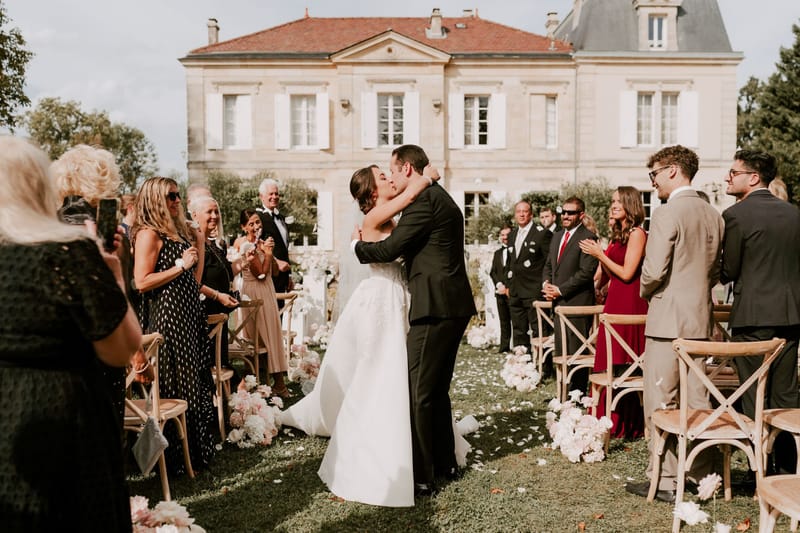 Bride and groom first kiss as guests toss petals on chateau lawn with blush and white floral aisle markers