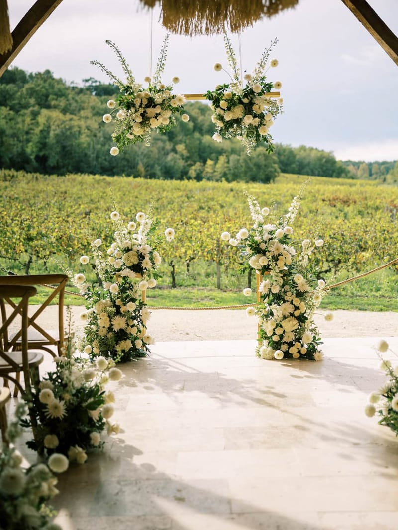 White dahlia and delphinium floral arch suspended from barn beams with matching column arrangements overlooking vineyard