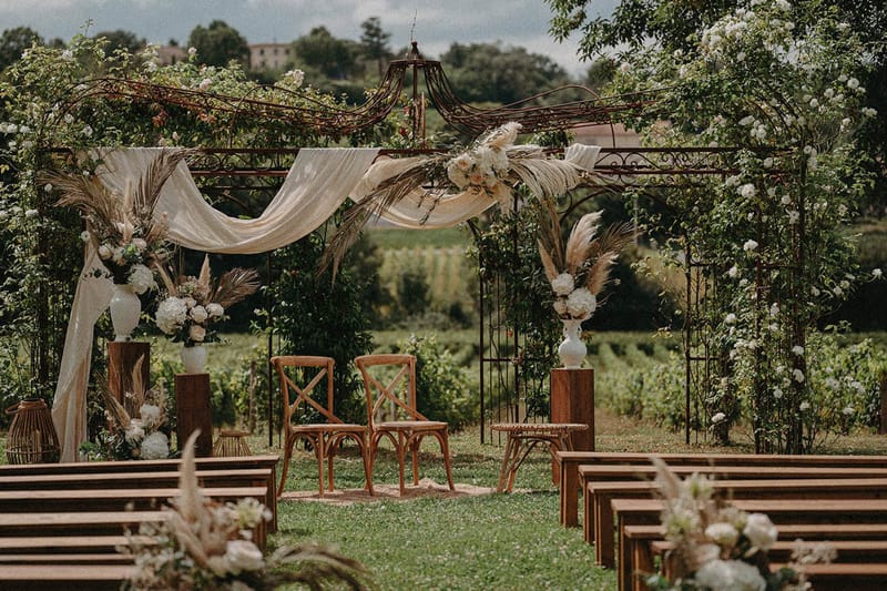 Outdoor ceremony setup with wrought-iron pergola draped in white roses, pampas grass, and wooden bench seating along viney...