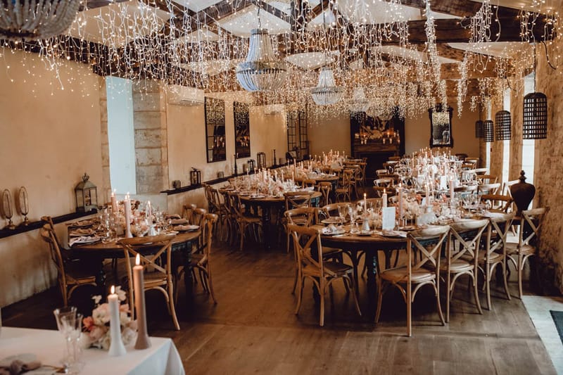Indoor reception room with fairy-light-draped ceiling beams, crystal chandeliers, and blush floral centerpieces on wood ta...
