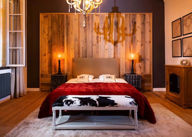 Guest bedroom in French chateau with taupe headboard, red velvet bedspread, reclaimed wood accent wall, and gold chandelier