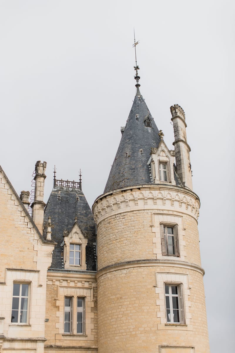Château exterior with circular tower, conical slate roof, pale golden stone, and adjacent mansard wing with decorative chimneys