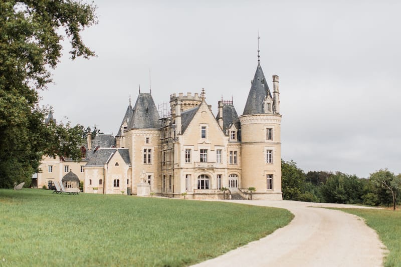 A wide exterior shot of a French château featuring pale golden limestone architecture with slate-grey conical turret roofs, crenellated battlements, and pointed dormer windows across multiple stories. The building includes a rounded tower on the right, ornate Gothic-Revival detailing, and a decorative stone statue mounted on the central facade. A curved gravel driveway leads toward the entrance, and sun loungers are visible on the left side of the grounds. Scaffolding is partially visible on the upper left section of the building, indicating ongoing restoration work. No people are present in the image. Potential venue feature image.