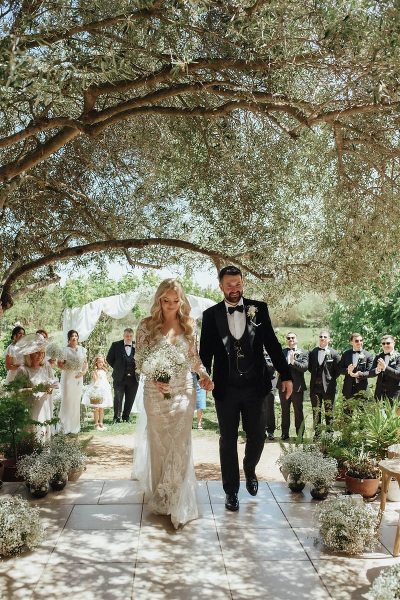 The bride and groom walk back up the aisle together following an outdoor ceremony, set on a tiled path beneath large, arching olive trees that create a natural canopy overhead. The bride wears a long-sleeved, fitted lace gown in ivory with intricate floral detailing and carries a large bouquet of white gypsophila (baby's breath); the groom wears a black tuxedo with bow tie, a three-piece suit with pocket watch chain, and a gypsophila boutonniere. The aisle is lined with large potted arrangements of white gypsophila, creating a consistent all-white floral theme throughout the ceremony space. In the background, approximately ten guests — several men in black tuxedos and a few women in white outfits — applaud the couple, with a white draped fabric arch visible at the far end; a small flower girl in blue is also visible among the guests. The shot is a wide, portrait-orientation image taken from ground level looking toward the couple as they approach the camera.