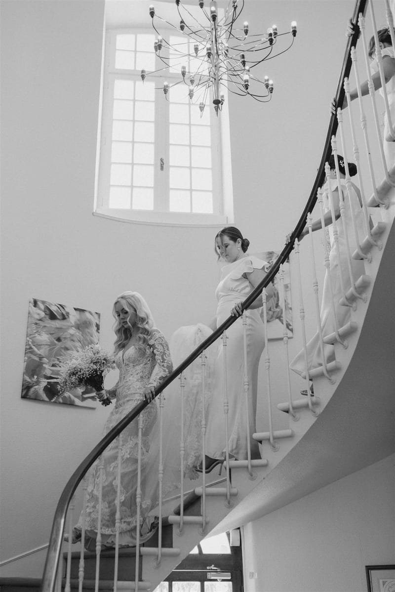 Black and white bride descending curved chateau staircase with bridesmaids, modern chandelier above