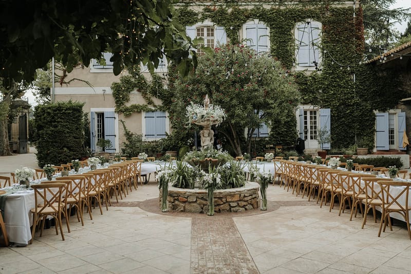 Outdoor reception with banquet tables around stone fountain in ivy-covered manor courtyard with fairy lights