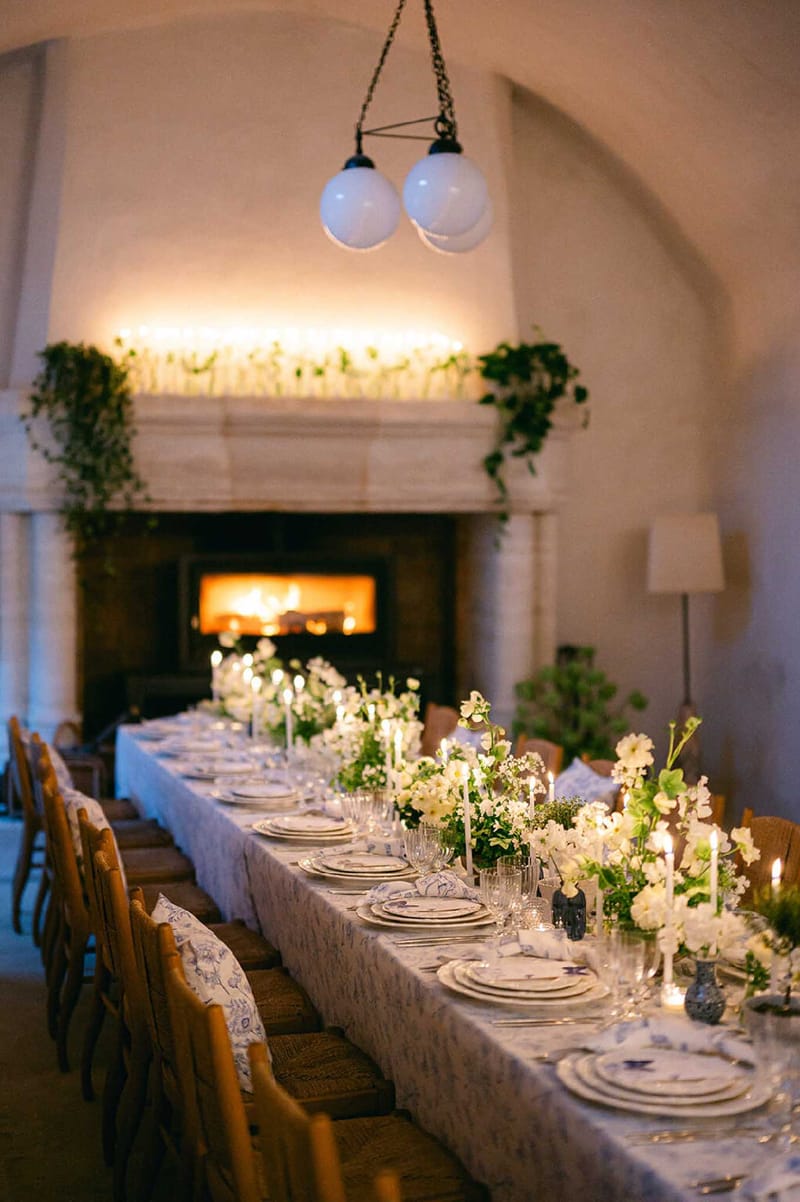 Long banquet table with blue-and-white toile cloth, white floral runner, and taper candles in a vaulted stone room