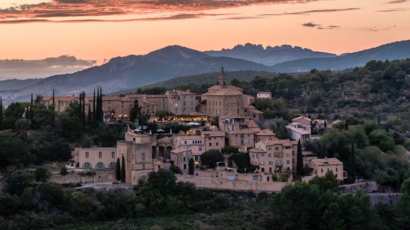 Wide aerial shot of a hilltop Provençal village or château complex photographed at dusk, with warm interior lights glowing from within the stone buildings. The architecture consists of multiple honey-toned limestone structures with terracotta-tiled roofs, a central tower with a bell or clock element, crenellated walls, and what appears to be a terrace area with white market umbrellas and string lights visible mid-complex. The surrounding landscape includes tall cypress trees interspersed among the buildings. A mountain range with jagged rocky peaks is visible in the background beneath an orange and pink dusk sky. No people are visible in the frame. Potential venue feature image.