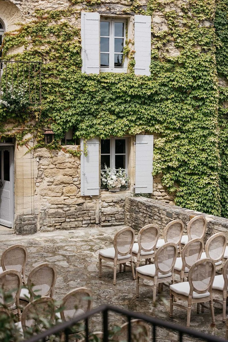 An outdoor ceremony setup in a stone courtyard in front of a French château or manor house facade with pale grey shuttered windows. Rows of French Louis-style chairs with cane backs and cream upholstered seat cushions are arranged in a curved seating configuration facing the building. A floral arrangement of white and cream blooms is displayed on the lower windowsill as a ceremony backdrop decoration. The overall decor palette is neutral — natural wood tones, cream, and soft grey — consistent with a classic French country styling theme. The image is a wide shot capturing both the seating arrangement and the architectural facade. Potential venue feature image.