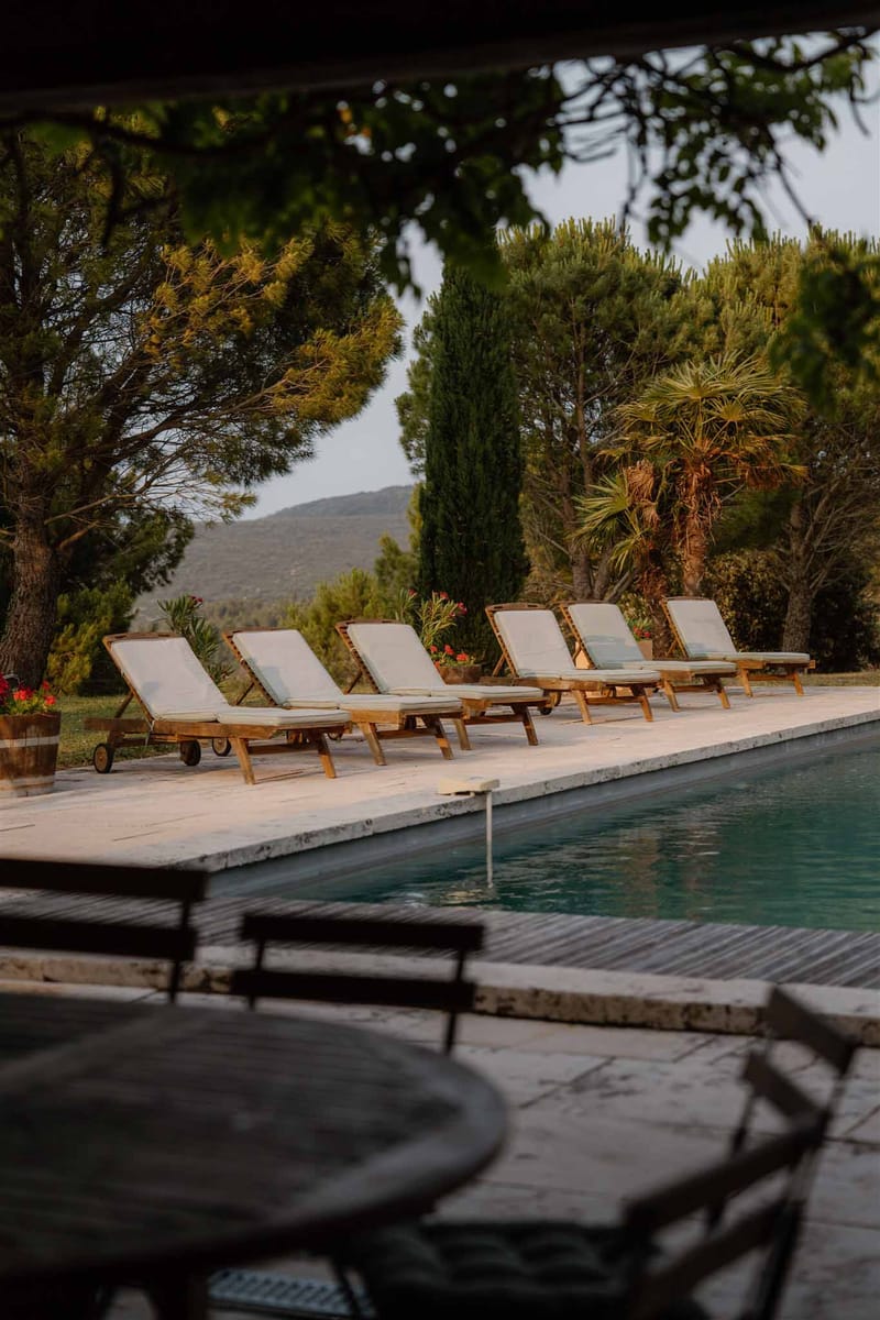 An outdoor pool area at a French countryside venue, photographed at golden hour with warm, low light across the stone pool deck. Six teak sun loungers with off-white cushions are lined up along one side of a rectangular lap pool with a light stone coping edge. In the foreground, a dark metal bistro table and chairs sit in a shaded covered area. Red flowering plants in terracotta pots are visible along the poolside. The background features a view of rolling hills and a mix of cypress trees, pine trees, and a palm tree. No people are present in the image. Wide shot framing the full pool terrace from a slightly elevated, shaded vantage point. Potential venue feature image.