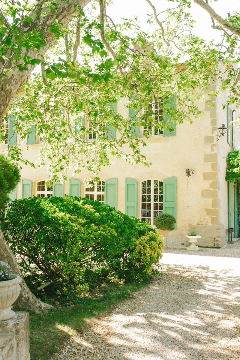 Cream limestone mas with sage green shutters, arched windows, and stone urn planters on gravel forecourt