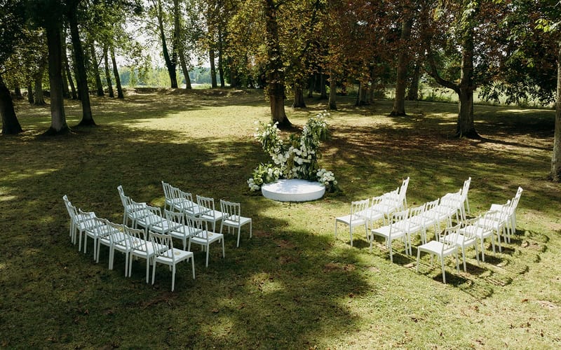 An outdoor wedding ceremony setup photographed from a slightly elevated wide-angle perspective, showing a wooded parkland setting with tall mature trees and dappled sunlight. Two sections of white Napoléon-style chairs are arranged in curved rows facing a circular white platform altar at the center, which is adorned with a large floral arrangement of white flowers, green foliage, and trailing greenery. The seating configuration accommodates approximately 40–50 guests across both sides. The overall decor palette is white and green, with a modern-classic styling approach using clean lines and a minimal color scheme. No people are present; the space is set up and awaiting the ceremony.