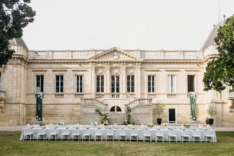 Long banquet tables with white and sage green linens set on lawn before a classical French chateau with double staircase