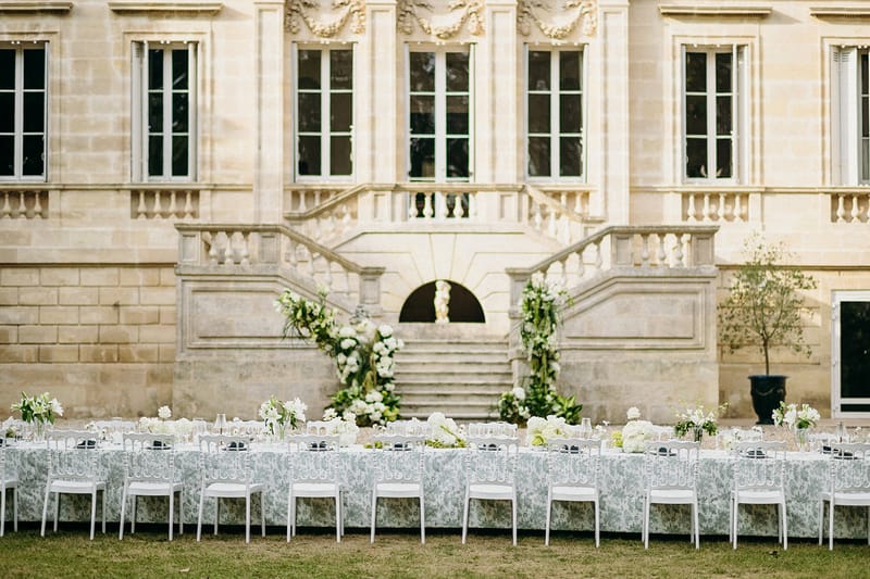 Blue toile table with ghost chairs and white floral staircase arrangements at classical chateau courtyard
