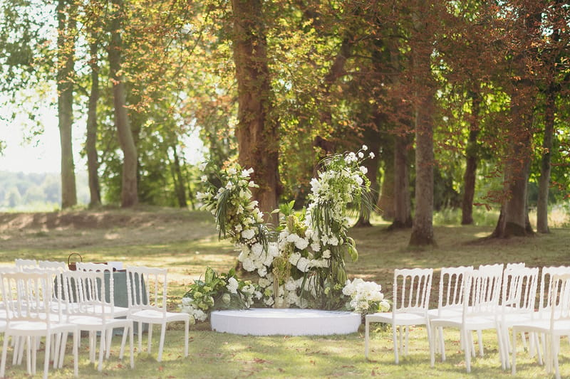 Outdoor circular ceremony setup with white chairs and freestanding white hydrangea and lily floral installation