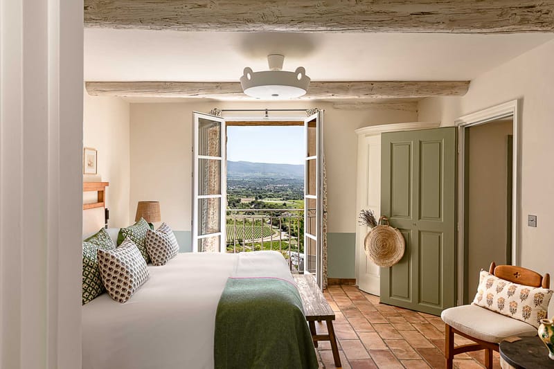 This image shows a hotel or venue bedroom interior, not a wedding photograph. The room features a double bed dressed in white linens with a forest green throw blanket and an assortment of block-print patterned cushions in green, white, and brown tones. The decor follows a Provençal style with terracotta tile flooring, exposed whitewashed wooden ceiling beams, sage green painted armoire and doors, a rattan lampshade, and a small wooden bench at the foot of the bed. French doors open onto a wrought-iron balcony revealing a wide landscape view of vineyards and countryside below. A wicker round bag and a small bunch of dried lavender hang on the armoire. The shot is a wide interior perspective taken from the doorway. Potential venue feature image.