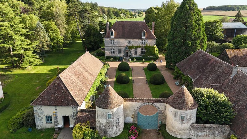 An aerial drone shot of a French manor house and its grounds, captured in full daylight. The property features a stone manor with a steeply pitched terracotta-tiled roof, flanked by outbuildings and a gatehouse entrance with two round stone towers topped with conical roofs and a pale blue arched gate. The formal forecourt between the gate and the manor entrance is laid out with a gravel driveway lined by symmetrically placed clipped topiary spheres and ground-level lanterns placed along the path. A small group of two or three people can be seen near the manor's front entrance, suggesting wedding activity in progress. Pink flowering shrubs are visible near the gatehouse towers. Potential venue feature image.