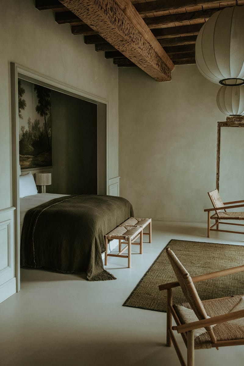 This image shows a hotel or château bedroom interior with no wedding-related activity visible. The room features exposed dark wood ceiling beams, smooth pale plaster walls with white panelling details, and a polished concrete floor. The sleeping area is set into an alcove painted in deep olive green, with a large dark olive linen bedspread with fringe detail, white pillows, a small bedside lamp, and a large landscape oil painting as a headboard backdrop. A woven rattan bench sits at the foot of the bed, two natural wood and woven rush lounge chairs are positioned in the main room alongside a dark olive jute area rug, and an oversized white spherical paper pendant lamp hangs from the ceiling. The styling is minimal and earthy with a muted olive, natural wood, and off-white palette. Wide interior shot. Potential venue feature image.