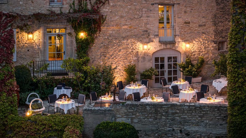 Evening reception setup on stone chateau terrace with round tables, white linens, candlelit centerpieces, and amber upligh...