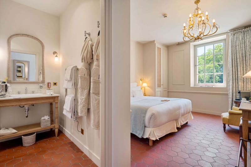 Interior shot of a hotel or château guest room shown as a split view, with a vanity/bathroom area on the left and the bedroom on the right. The bathroom side features a natural oak washstand with a white basin, an ornate silver-gilt arched mirror, a wall sconce with warm bulb lighting, and white towels and robes hung on wall hooks, with a champagne bucket visible on the counter. The bedroom side features a double bed dressed in white linens with a pale blue throw, terracotta hexagonal floor tiles, cream-painted panelled walls, a gold multi-arm chandelier, sage green patterned curtains framing a large window, a small bedside lamp, and a yellow armchair with a writing desk. The overall décor palette is cream, terracotta, and muted gold in a classic French country house style. Wide interior shot. Potential venue feature image.
