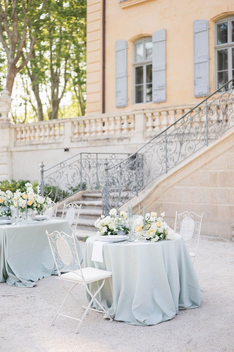 Round tables in sage linen with ivory rose centerpieces white bistro chairs before ochre chateau with scrollwork staircase