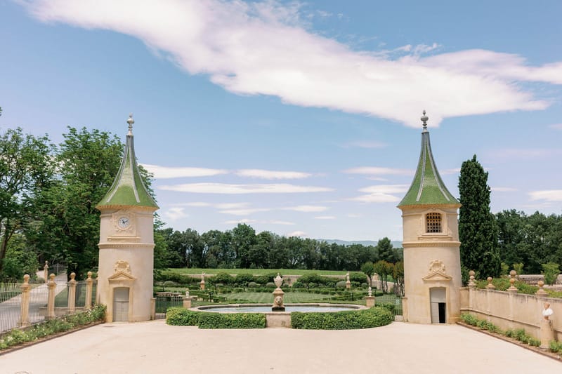 French chateau entrance with twin stone pavilion towers, formal boxwood garden, circular fountain, and vineyard beyond