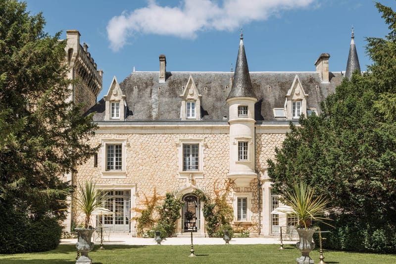 French chateau exterior with golden limestone facade, slate mansard roof, round turret, iron gate, and stone urns