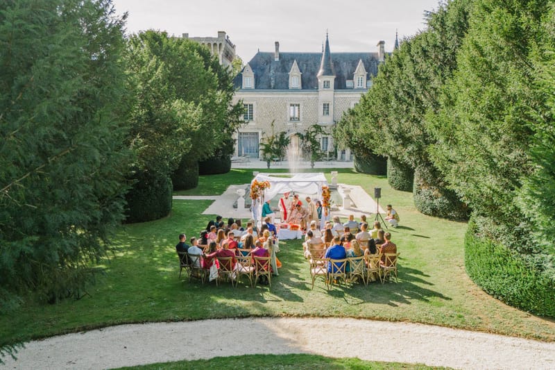 Elevated view of Hindu wedding ceremony under white mandap with orange florals in French chateau garden with seated guests