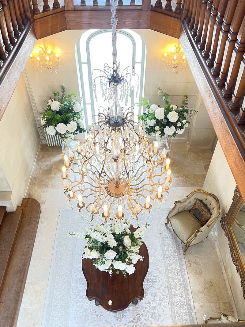 An overhead shot looking down into the grand entrance hall of a château or manor house, decorated for a wedding. The space features a large multi-tiered gold crystal chandelier with lit candle-style bulbs hanging in a double-height stairwell flanked by dark wood balustrades. Three floral arrangements of white hydrangeas, white roses, and lush green foliage are positioned throughout the hall: two large urns flanking a tall arched window at the back, and one arrangement on a dark wood console table directly below the chandelier. A light-colored ornate rug covers the stone tile floor, and a gold-framed upholstered armchair is visible in the lower right corner alongside what appears to be a gilded mirror. The decor palette is white and green with gold accents, in a classic French interior style. Potential venue feature image.