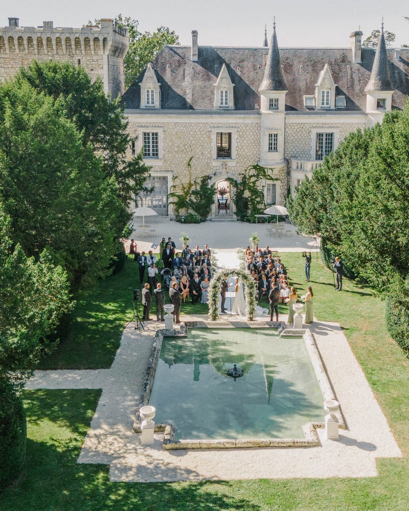 Aerial ceremony in chateau parterre garden eighty guests facing floral arch reflecting pool and turreted stone facade