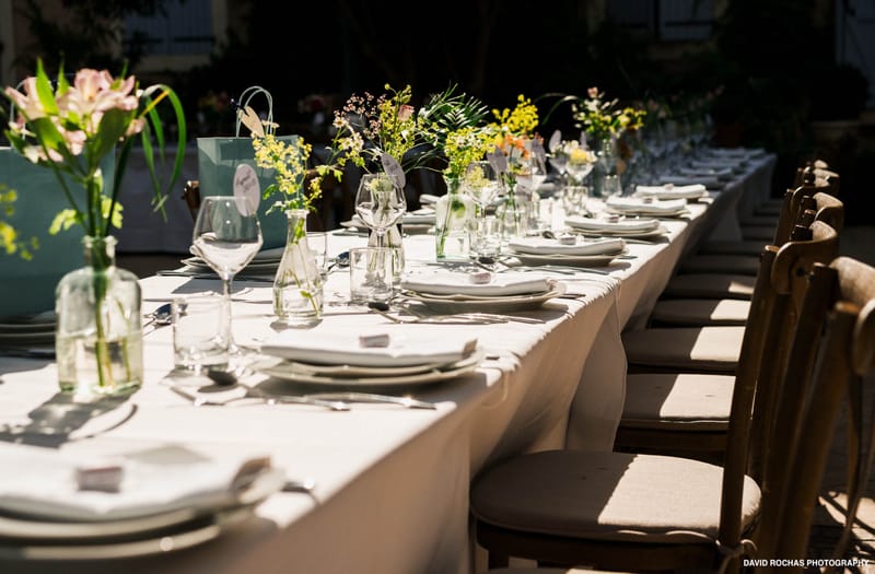 A detail shot of a long outdoor wedding reception table set for a large number of guests, shot from a low angle along the length of the table. The table is covered with a white linen cloth and laid with white ceramic plates, folded white napkins, silver cutlery, and clear wine glasses. Centerpieces consist of mismatched clear glass bottles and bud vases holding loosely arranged stems of yellow oncidium orchids, pink alstroemeria, and small wildflower-style blooms in yellow, pink, and green, creating an informal, garden-style aesthetic. Small place cards or favor tags are visible at each setting. Light teal gift bags are placed at intervals along the table. The chairs are natural wood cross-back style with beige cushioned seats. Natural sunlight casts dappled shadows across the table, suggesting the setting is outdoors in a shaded courtyard or garden area.