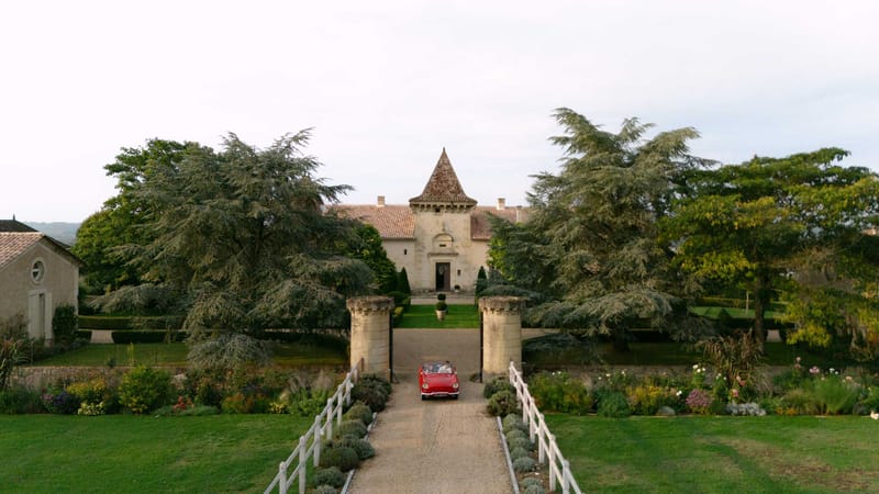 Vintage red convertible driving up gravel driveway toward French chateau with stone turret and flower-lined path