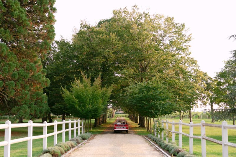 Vintage red Citroen driving down tree-lined gravel driveway with white fencing and vineyard views at French wine estate