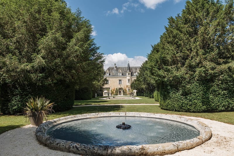 French chateau with slate mansard roof and turret viewed from formal garden with stone fountain and clipped hedges