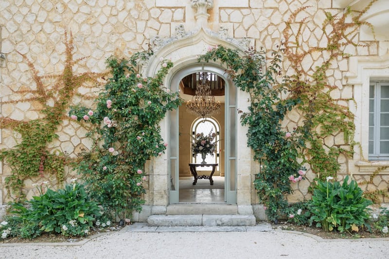Chateau entrance with climbing roses, blue-grey doors, crystal chandelier, and blush rose arrangement in stone urn