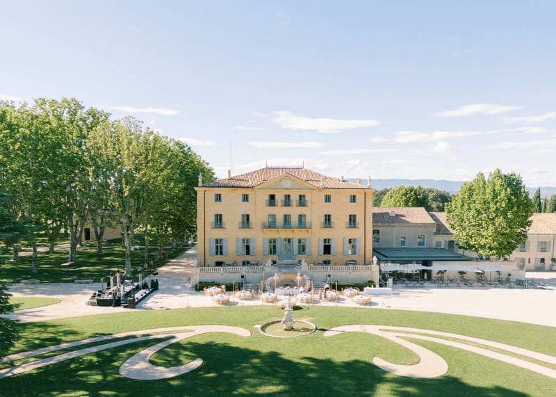 Aerial view of a yellow-ochre bastide chateau courtyard set with round reception tables around a stone fountain