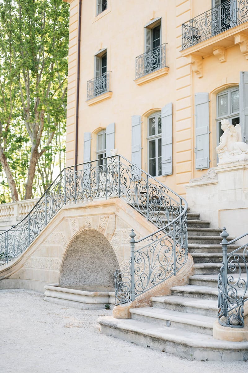 Grand double staircase with wrought-iron scrollwork leading to peach-ochre chateau facade with blue shuttered arched windows