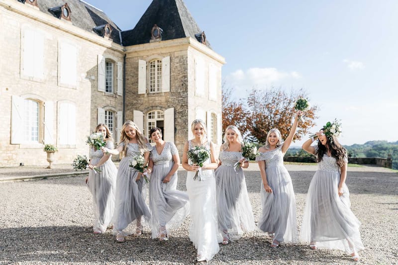Bride in ivory lace gown walking with six bridesmaids in silver-grey tulle dresses on chateau forecourt